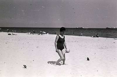 A woman in a black one-piece swimsuit walks along a sandy beach, clutching a beach ball. Several figures are visible in the b...
