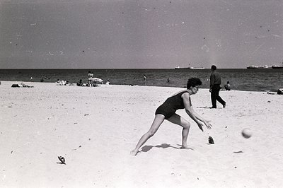 A woman in a one-piece swimsuit leaps forward to catch a ball on a sandy beach. Background shows other beachgoers and ships a...