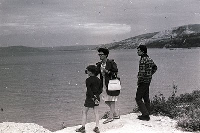 A family - woman, man, and child - stand overlooking a vast seascape. The woman wears a dress and carries a handbag; the man,...