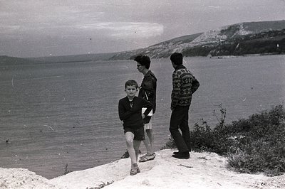 A young boy runs along a rocky clifftop overlooking a wide expanse of sea. Two other figures, one in a jacket and one in a pa...