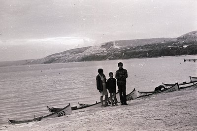 Seaside scene featuring three figures—two boys and a man—standing among rowboats on a sandy beach. A hillside with sparse veg...