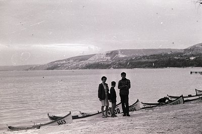 Three figures stand on a beach beside an array of small, numbered boats, likely canoes. The background features a gently slop...