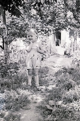 A young child, possibly a toddler, stands on a dirt path within a leafy garden. They are wearing a floral-print romper and ha...