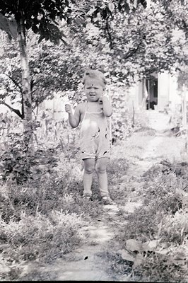A young child, possibly toddler, stands in a garden path, exhibiting distress – tears and a hand to the face. The child wears...