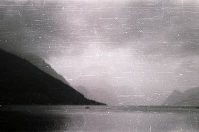 Striking monochrome landscape featuring a dark, forested mountain slope rising from a body of water. Distant, hazy peaks are ...