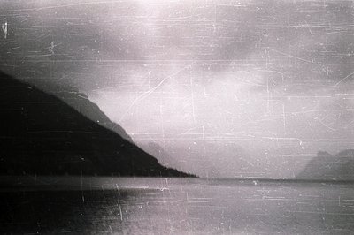 Striking monochromatic landscape showcasing a placid lake reflecting a clouded sky and a dark, steep shoreline. Visible film ...
