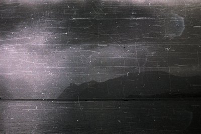 Seascape featuring a dark body of water and distant, silhouetted mountains under a cloudy sky. Visible scratches & aging mark...