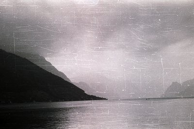 A panoramic coastal view; dark, rugged mountains rise from a calm body of water, likely a fjord or sea inlet. Visible grain a...