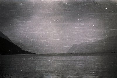 A black and white landscape shows a body of water extending to a distant shore defined by mountains. The image exhibits signi...