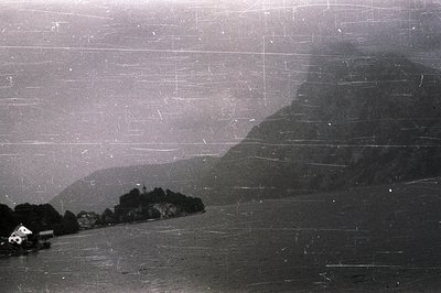 A hazy, grayscale view of a fjord landscape with a large mountain rising on the right. A small, white building sits on the sh...