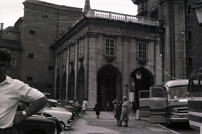 Classic architectural facade with arched portico and balcony, framed by surrounding buildings. Several vintage cars and a bus...