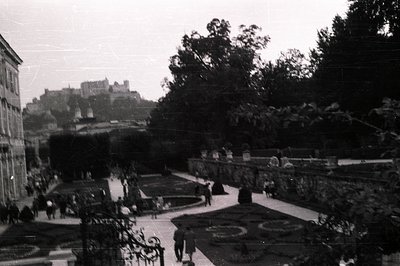 Formal garden featuring manicured hedges, patterned flowerbeds, and stone pathways. A distant castle silhouette dominates the...