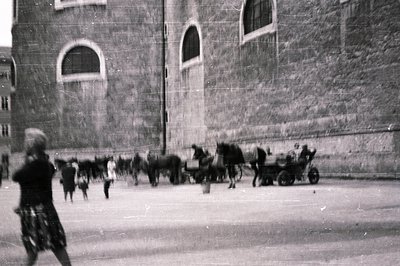 A bustling plaza with a stone building façade featuring arched windows. A horse-drawn cart and several horses are central. A ...