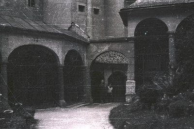 Series of arched passageways form a courtyard with stone paving, leading to a distant arched entrance. A person stands near t...