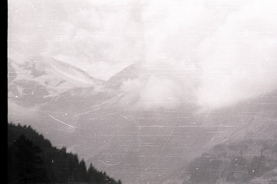 A black and white landscape depicts snow-covered alpine peaks shrouded in mist. Dense evergreen forest occupies the lower fra...