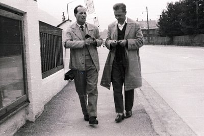 Two men in overcoats walk along a sidewalk, appearing deep in conversation. Architectural details suggest a residential area,...