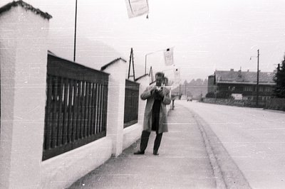 B&W street scene; a man in a long coat stands photographing with a vintage camera. Whitewashed wall & gate dominate left. Spa...