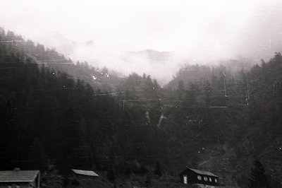 Monochrome image depicts a dense, coniferous forest valley shrouded in mist. Several small buildings are visible amongst the ...