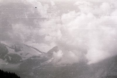 Dramatic, monochromatic view of a mountainous landscape obscured by thick, swirling clouds. Visible peaks suggest alpine terr...
