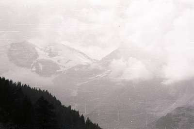 Monochrome landscape depicting a mountainous, forested region shrouded in mist and fog. Visible slopes suggest winter conditi...
