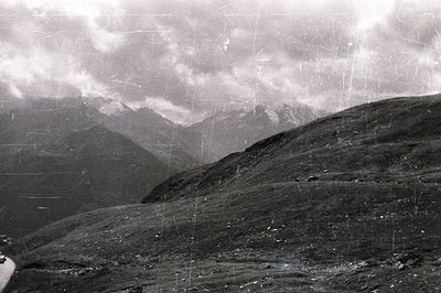 Dramatic, monochrome landscape showcasing a rugged, alpine terrain under a heavy, overcast sky. A winding road is visible in ...