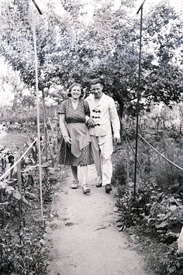 A couple strolls along a gravel path flanked by lush foliage, showcasing 1960s fashion. The woman wears a patterned dress; th...