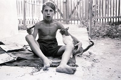 A barefoot boy, shirtless and wearing dark shorts, sits outdoors with a tabby cat perched on his lap. He holds a small object...