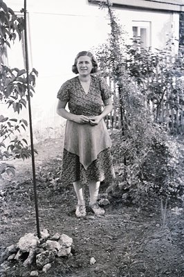 A woman stands in a garden with a stone wall and lush foliage in the background. She wears a patterned, knee-length dress wit...