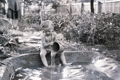 A young girl sits in a shallow, stone-lined pool, playfully pouring water from a bucket. Lush foliage fills the background, w...