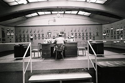 A man sits at a control panel in a large, curved room, likely an industrial or scientific facility. Numerous gauges, dials, a...
