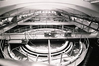 Industrial scene: circular water treatment basin with tiered platforms and railings. Visible piping and machinery suggest a f...