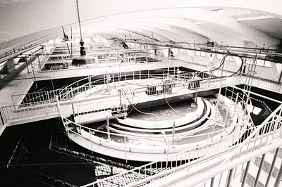 Elevated view of an indoor, tiered fountain structure. White railings and walkways frame dark, reflective pools arranged in c...