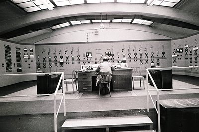 Man seated at a control panel densely populated with switches and meters within a large, high-ceilinged room. Likely a teleco...