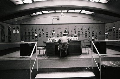 A technician sits at a control console within a large, industrial room. Numerous panel meters and displays cover the walls, s...