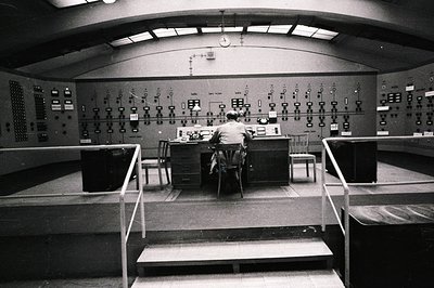 A technician sits at a control panel within a stark, elevated room. Numerous gauges, switches, and indicator lights cover the...