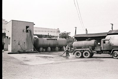 Industrial scene: Large fuel storage tanks dominate the view, with a tanker truck parked beside them, attended by a worker. S...