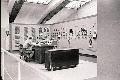 Control room interior, likely an industrial facility. Two men seated at a large console monitoring complex control panels fil...
