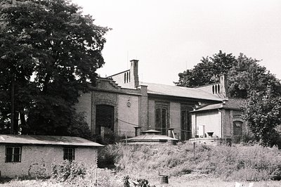 Dilapidated brick building with arched windows and chimney stacks, partially obscured by dense trees. Adjacent smaller struct...