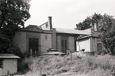 A brick building with elaborate detailing, including a round turret and decorative chimneys, sits amidst overgrown vegetation...
