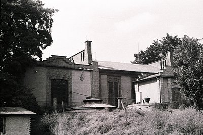 A two-story brick building with arched windows and decorative stonework, set within overgrown vegetation. Note the prominent ...