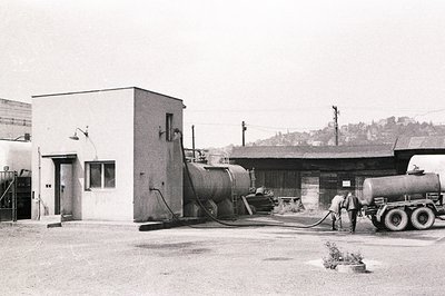 Industrial landscape featuring large storage tanks and a utilitarian building with a prominent chimney. Appears to be a proce...