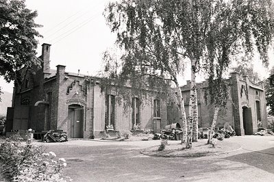 A view of a brick industrial building with distinctive crenellations and arched doorways. Adjacent workshops are visible, wit...