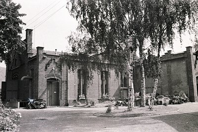 A brick building with arched entry and decorative detailing faces a tree-lined street. A vintage car is parked nearby. Likely...