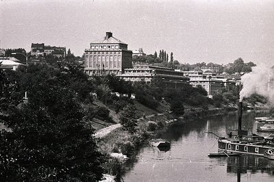 A large industrial building with a central cupola dominates a riverside scene. A steamboat emits steam on the waterway, sugge...