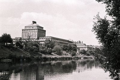 Architectural view of a grand, multi-story building with a prominent central tower. Situated on an elevated riverbank, it fea...