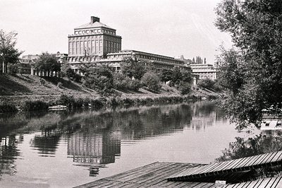 A grand, neoclassical building with a central cupola dominates this landscape, reflected in a calm body of water. Likely a pu...
