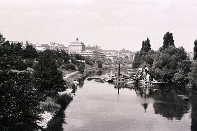 A wide river reflects a large, ornate building with a central tower and surrounding smaller structures. A dredging barge occu...