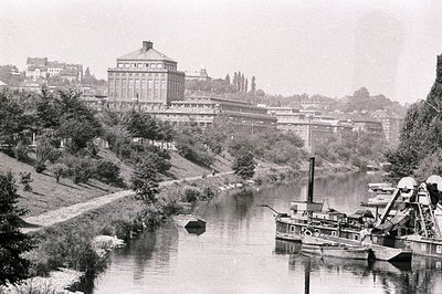 A long view of a river, likely the Rhine, with a large industrial building complex dominating the hillside. A dredging boat i...
