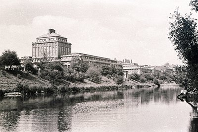 Grand, neoclassical building complex sits atop a grassy bank overlooking a river. Multiple wings extend from a central, promi...