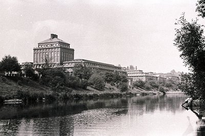 Grand public building with a central tower and substantial wings, situated on a rise overlooking a tranquil river. Architectu...